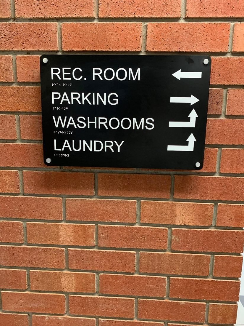 Black directional sign on a brick wall pointing to the rec room, parking, washrooms and laundry